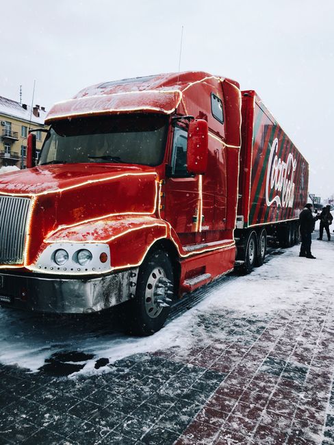 A large red Coca-Cola delivery truck parked on a snow-covered pavement outdoors during winter, with snow accumulating on its roof and front. The truck features a prominent Coca-Cola logo on the side, 