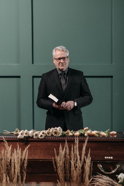 A well-dressed man with grey hair and glasses stands behind a dark wooden table, holding a small book or card. In front of him, on the table, are several floral arrangements consisting of white and pi