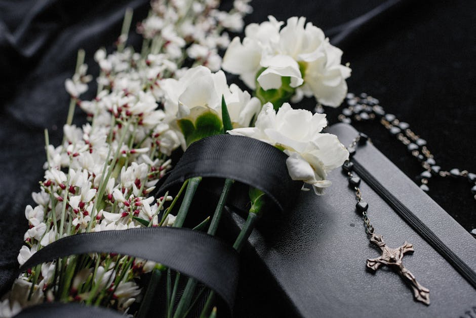 A close-up of white carnations and small white flowers wrapped with black satin ribbons, arranged on a dark surface. Next to the flowers, there is a black book or keepsake box with a silver chain and 