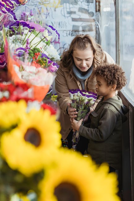 A young woman and a small boy are inside a floral shop near a large window, engaged in a floral transaction. The woman, with wavy light brown hair, is gently leaning towards the boy, offering him a sm